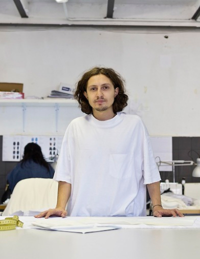 A person with long hair and a white T-shirt standing behind a table with some papers